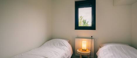 A small, single-room accommodation with two single beds, a small table with a lamp, and a window with a view of greenery.