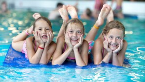 Three children lying on a blue floating mat in a swimming pool.