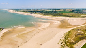 Ein Luftbild von einem Strand mit klarem Wasser, Sandküste und im Hintergrund einer Mischung aus Feldern und Gebäuden.