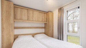 Ein Schlafzimmer mit einem hölzernen Kopfende, zwei Betten, einem Fenster mit Vorhängen und Blick auf Bäume.