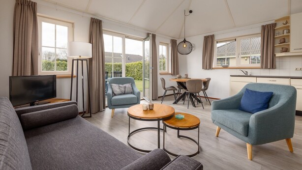 Een ruime woonkamer met een bank, fauteuil en een televisie. Er is een eethoek met een tafel en stoelen, en een keuken op de achtergrond.