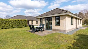 Een tweelaags huis met een terras, een eettafel met stoelen en een haag.