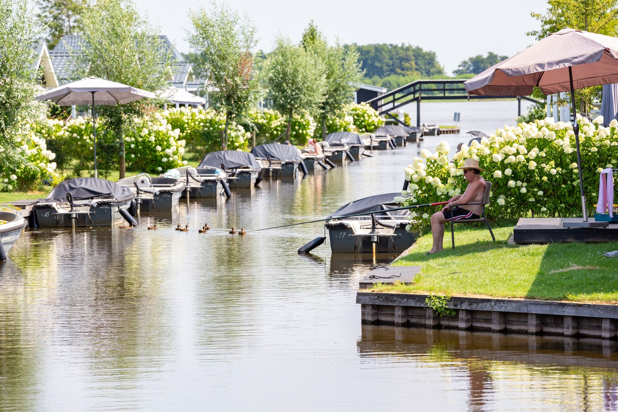 Een oeverlandschap met boten en zonneschermen, een persoon zittend op een steiger, en een achtergrond van bomen en bloemen.
