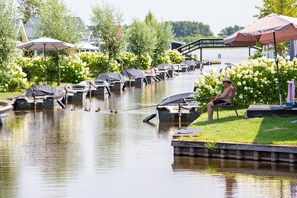 Une zone riveraine avec des bateaux et des auvents, une personne assise sur un quai, et un paysage en arrière-plan composé d’arbres et de fleurs.
