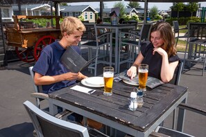 Dos personas sentadas en una mesa exterior con una mesa de madera, sillas de metal y una cerveza.