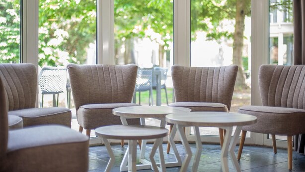 Een zithoek met gestoffeerde stoelen en kleine ronde tafels, grote ramen met uitzicht op groen.