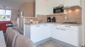 A compact kitchen with white cabinets, a microwave, a gas stove, and a coffee maker. There is a window providing natural light and a seating area with a red couch and a table.