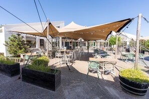Ein Terrassenbereich im Freien mit mehreren Tischen und Stühlen, beschattet durch große Sonnensegel, umgeben von Pflanzkästen mit Grünpflanzen.