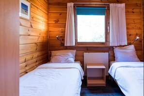 Ein Holzhüttenraum mit zwei Betten, einem Vorhangfenster, einem Heizkörper und einem kleinen Tisch.