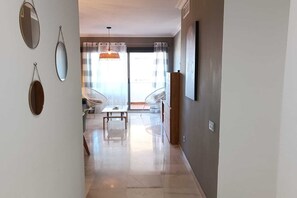A hallway leading to a living area with a sofa, a dining table, and a chair. There are two hanging mirrors and a pendant light.