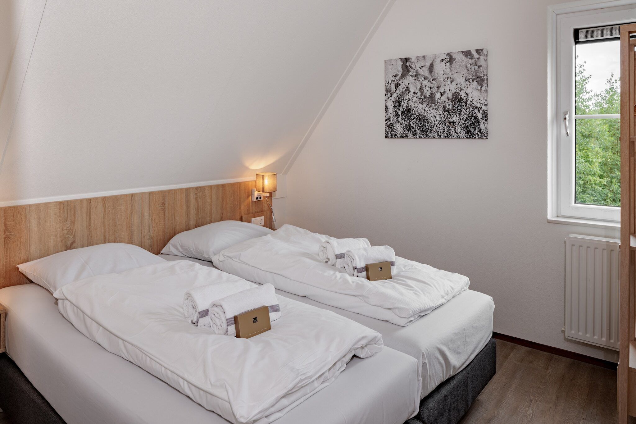 Une chambre d’hôtel avec deux lits, une tête de lit en bois, une lampe fixée au mur et une fenêtre donnant sur un paysage verdoyant.