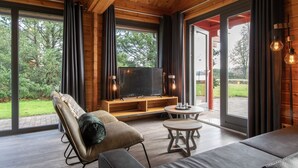 Een moderne woonkamer met een grote televisie, houten meubels en grote ramen met uitzicht op het groen.
