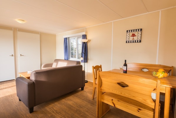A room with a brown sofa, a wooden dining table with a bottle and fruit, and a window with curtains.