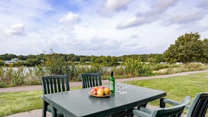 A table with a basket of fruit, a bottle, and glasses, set outdoors with a view of a lake and greenery. - Nature Stay in Hunzedal (Borger)