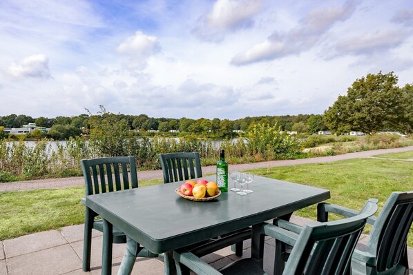 Una mesa con una cesta de fruta, una botella y vasos, al aire libre con vista a un lago y vegetación.