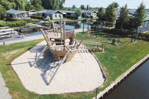 A playground with a swing set and a seesaw, surrounded by a sandy area and a water body. - Nice chalet with ac and sloop (Giethoorn)