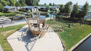 Une aire de jeux avec des balançoires et un toboggan, entourée d’une zone sablonneuse et d’un plan d’eau.