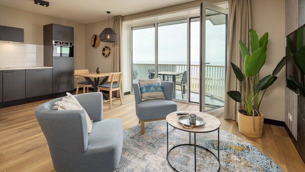 A modern living room with a sofa, armchair, and coffee table, a dining area with a table and chairs, a kitchen with grey cabinets, and a balcony with a plant.
