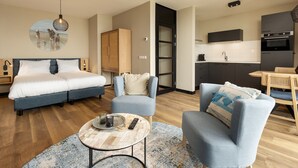 A modern hotel room with a double bed, a sofa, a coffee table, and a kitchen area.