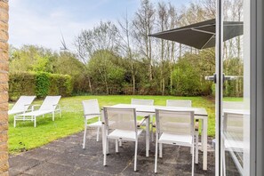 An outdoor seating area with white chairs and tables, a glass door, and a view of a grassy garden with trees and a wind turbine in the background.