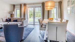 A modern living room with a sofa, a dining table with chairs, a blue rug, and a large window with curtains. - Villa in Banjaard near Sandy North Sea Beach (Kamperland)
