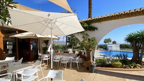 An outdoor patio area with white plastic chairs and tables, a large umbrella, and a view of a swimming pool and palm trees.