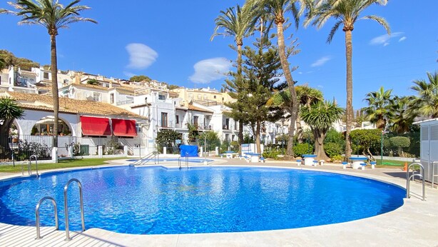 A swimming pool with a tiled deck, surrounded by palm trees and buildings with white facades and red awnings.