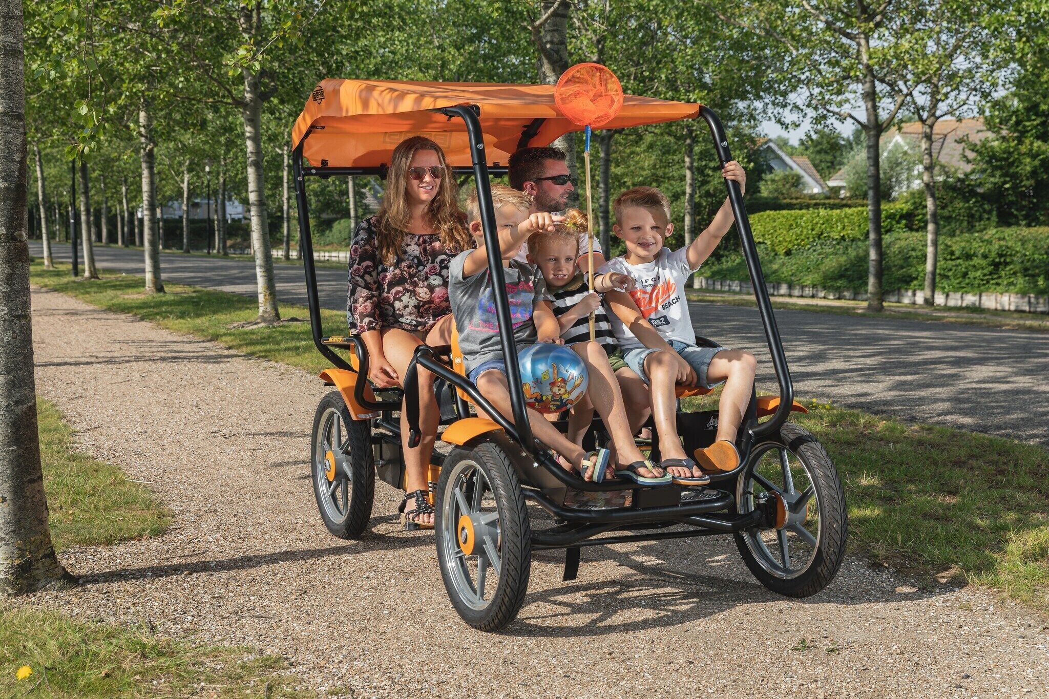 Una familia viaja por un camino de grava bordeado de árboles en un vehículo pequeño, naranja y de tres ruedas con toldo.