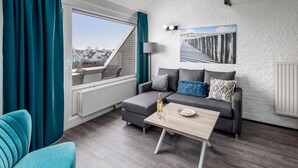 Un soggiorno moderno con un divano grigio, una poltrona color acquamarina e un tavolino in legno. Una finestra con tende mostra una vista esterna.