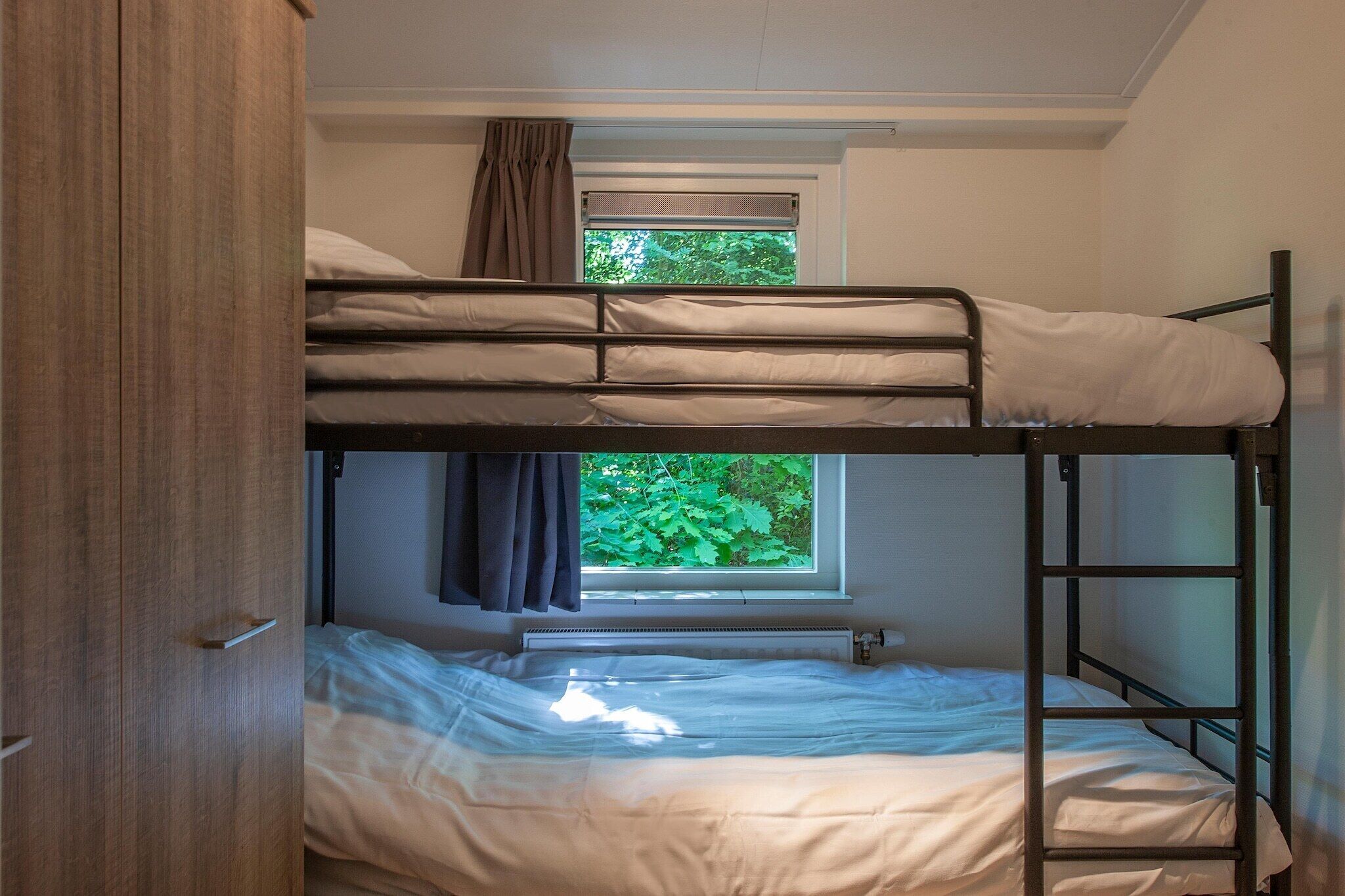 Een slaapkamer met een stapelbed, een raam, een radiator en een houten garderobe.
