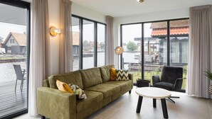 A modern living room with a green sofa, a round table, and large windows offering a view of a water body and buildings.