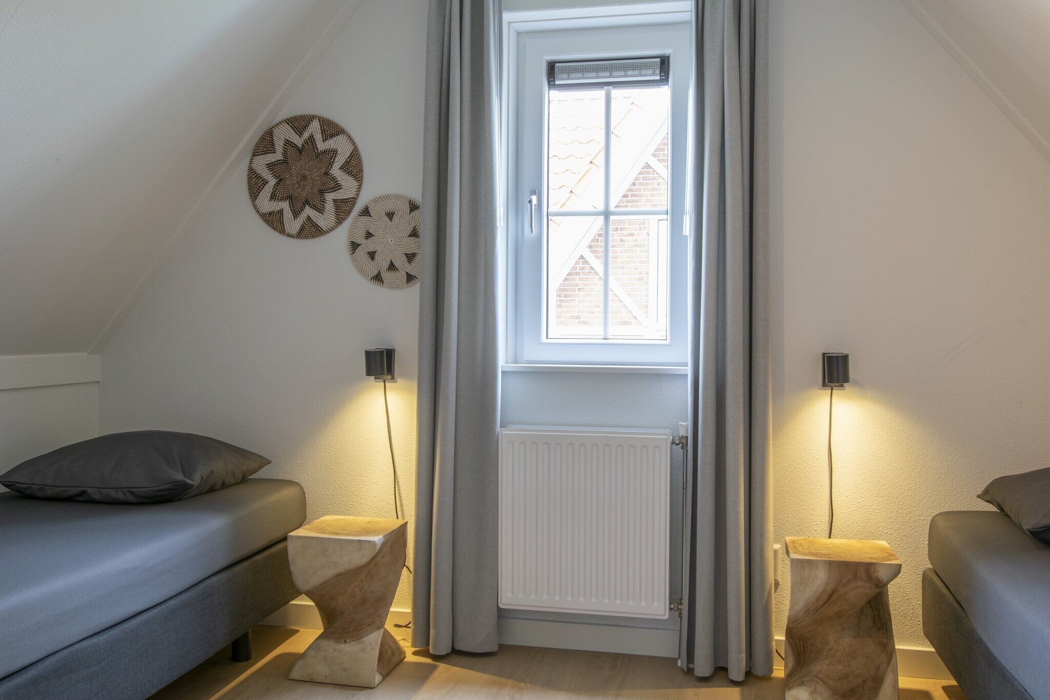 Ein gemütlicher Raum mit schrägen Wänden, einer weißen Tür mit Fenster, zwei Nachttischen mit Lampen und einem grauen Sofa.