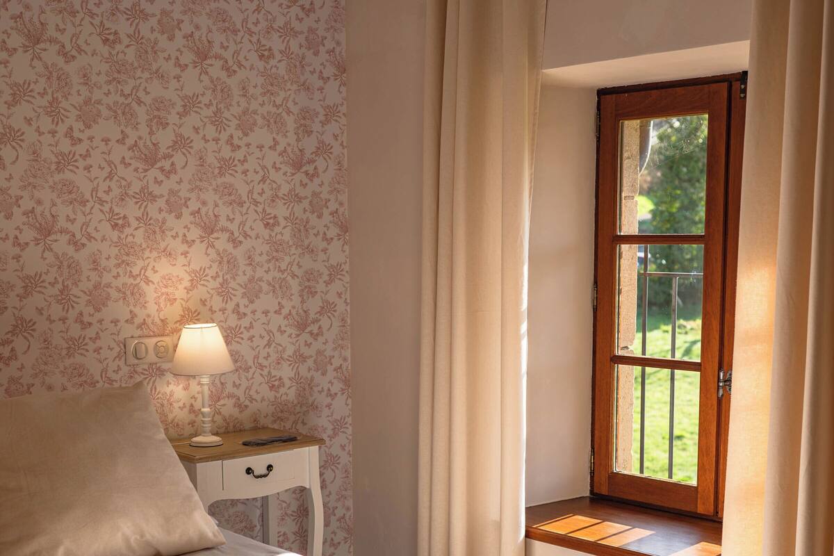Une chambre à coucher comprenant un lit, une table de chevet, une lampe, une fenêtre avec des rideaux et une vue sur la verdure à l’extérieur.