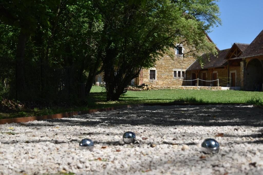 A courtyard with a gravel surface, a stone building with a tiled roof, and a large tree providing shade.