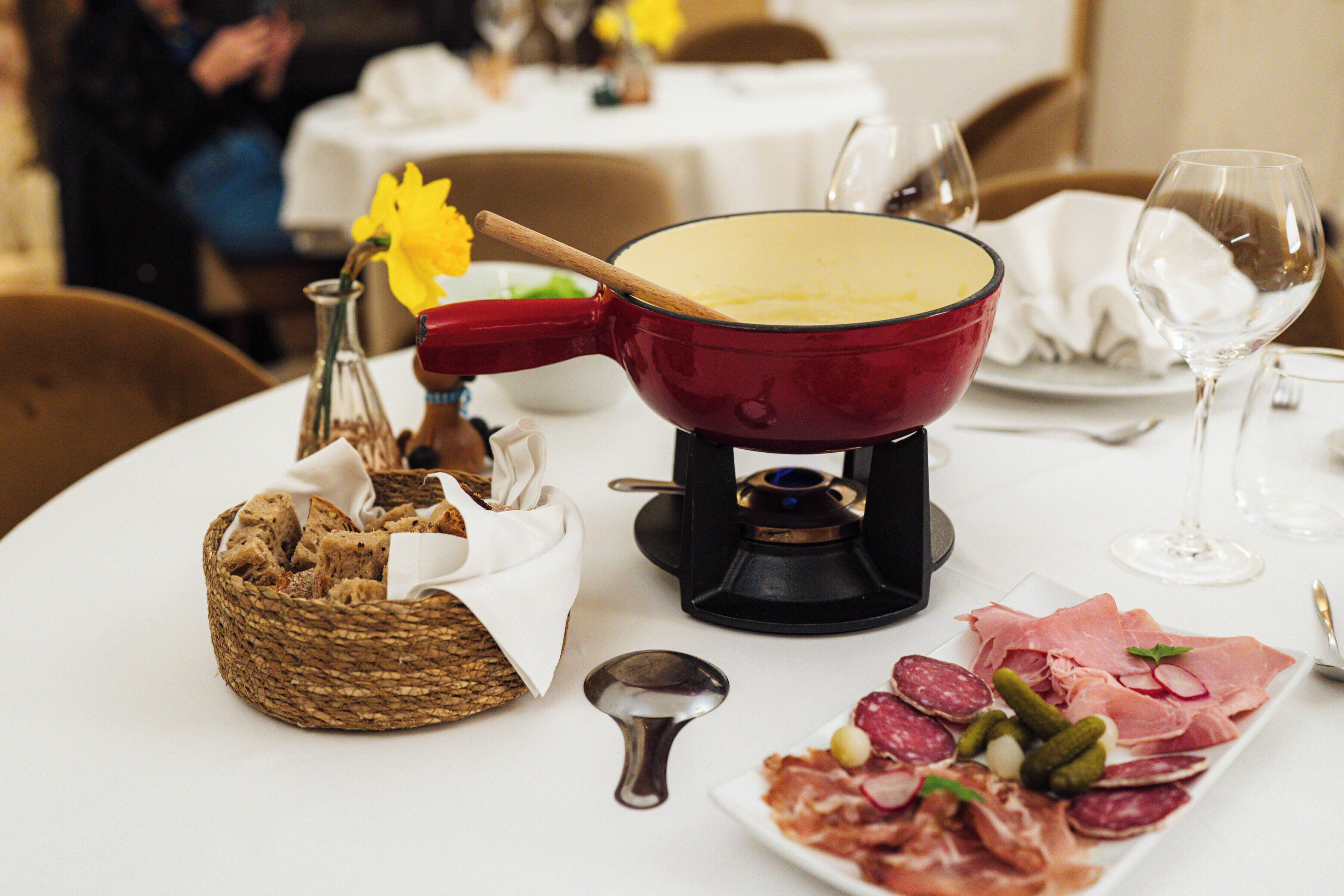 Une table à manger avec un caquelon à fondue, un panier de pain, une assiette de charcuterie et un verre à vin.