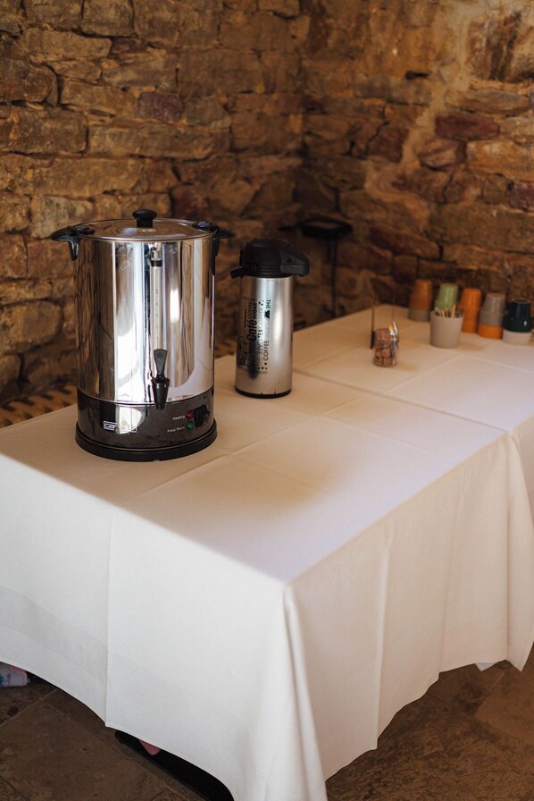Une table recouverte d’une nappe blanche, une casserole en acier inoxydable, un thermos et un mur en pierre en arrière-plan.