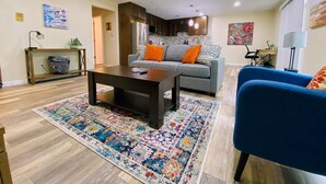 Un salon avec une table basse en bois foncé, un canapé gris agrémenté de coussins orange, un fauteuil bleu et un tapis aux couleurs vives.