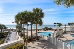 A pool area with a view of the ocean, palm trees, and a flag. - Sandy Shores Escape · Stunning beach views  *Not your grandma’s condo* (Miramar Beach)