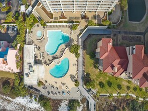 An aerial view of a resort with multiple swimming pools, a parking area, and a building with red-tiled roofs. - Sandy Shores Escape · Stunning beach views  *Not your grandma’s condo* (Miramar Beach)