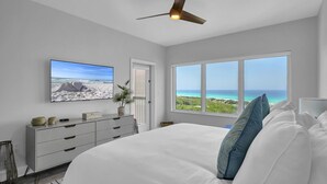 A bedroom with a large bed, a ceiling fan, a TV on the wall, and a window with a view of the ocean. - Sandy Shores Escape · Stunning beach views  *Not your grandma’s condo* (Miramar Beach)