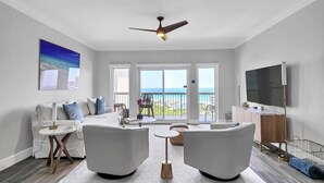 A modern living room with a white sofa, two armchairs, a coffee table, and a TV. There is a balcony with a view of the ocean. - Sandy Shores Escape · Stunning beach views  *Not your grandma’s condo* (Miramar Beach)