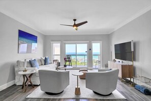 Ein modernes Wohnzimmer mit einem weißen Sofa, zwei Sesseln, einem Couchtisch und einem Fernseher. Ein Balkon bietet Blick aufs Meer.