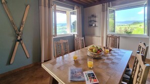 Un comedor con una mesa de madera, sillas, un frutero y una revista. Se observan ventanas con cortinas, un techo de madera y un estante en la pared.