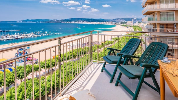 A balcony with two green chairs, a wooden table, and a view of a beach and marina. - Stylish Apartment in Palamós - Near Sea with Parking (Palamós)