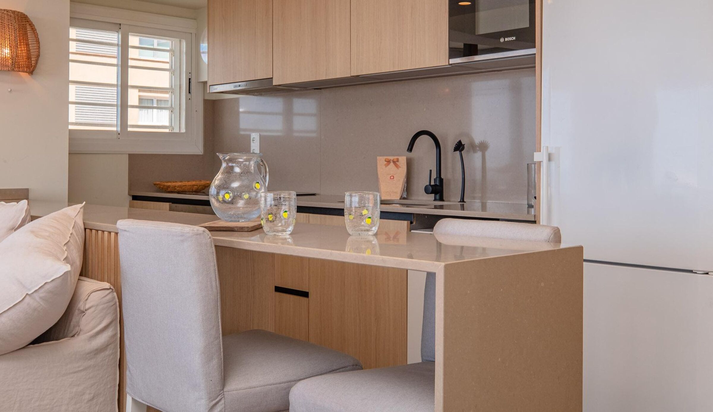 Uma kitchenette compacta com fogão embutido, pia e geladeira. Há um pequeno espaço de estar com um banco acolchoado e uma cadeira.