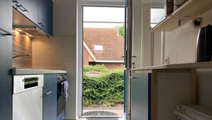 Una cocina compacta con electrodomésticos modernos, vista a un jardín a través de una puerta abierta y un pequeño espacio exterior con felpudo.