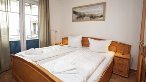 A hotel room with a double bed, a bedside table, a nightstand, a window with curtains, and a painting on the wall. - Die Ferienwohnung Austernperle im Haus am Kurpark in Baabe (Baabe)