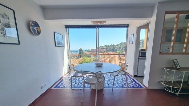 A dining area with a round table and chairs, a view through a glass door to the outside, and a kitchen area to the right. - Charming air-conditioned T2 near sea, 4 pers (Collioure)