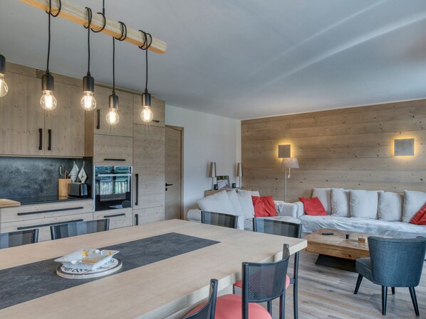 A modern, well-lit kitchen with wooden cabinets and pendant lighting, a dining area with a table and chairs, and a living space with a sofa and cushions.