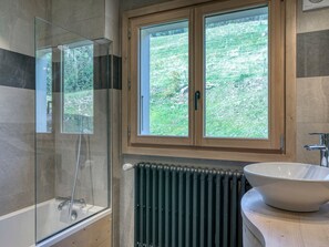 A modern bathroom with a glass-enclosed shower, a large window with a view of greenery, a white sink, and a radiator.